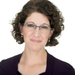 Headshot of Carol Smith, a middle-aged, curly haired white woman, wearing glasses and a wry smile.