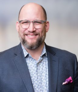 Headshot of Jeff Pass, a middle-aged, bald, bearded white male wearing glasses, a shirt, jacket and pocket square, with a squinting smile.