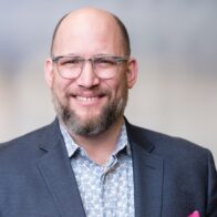 Headshot of Jeff Pass, a middle-aged, bald, bearded white male wearing glasses, a shirt, jacket and pocket square, with a squinting smile.