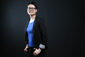 Regina Lam, woman of Asian descent wearing black rimmed glasses, black blazer over a bright blue shirt, smiling