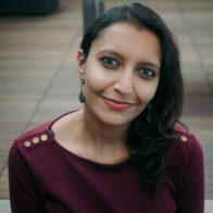 Woman of Indian descent, smiling up at the camera, wearing a maroon red top, black hair swept over the left shoulder