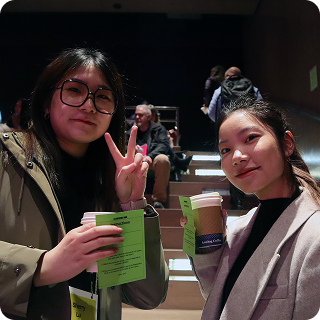 Two women facing the camera, holding drinks. They are in a lecture hall, and one is making a peace sign toward the camera.
