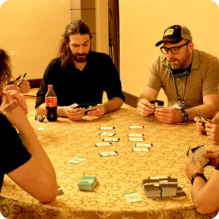 People sitting around a table playing a card game. Two men are seen on the far side of the table, looking at their cards in their hands.