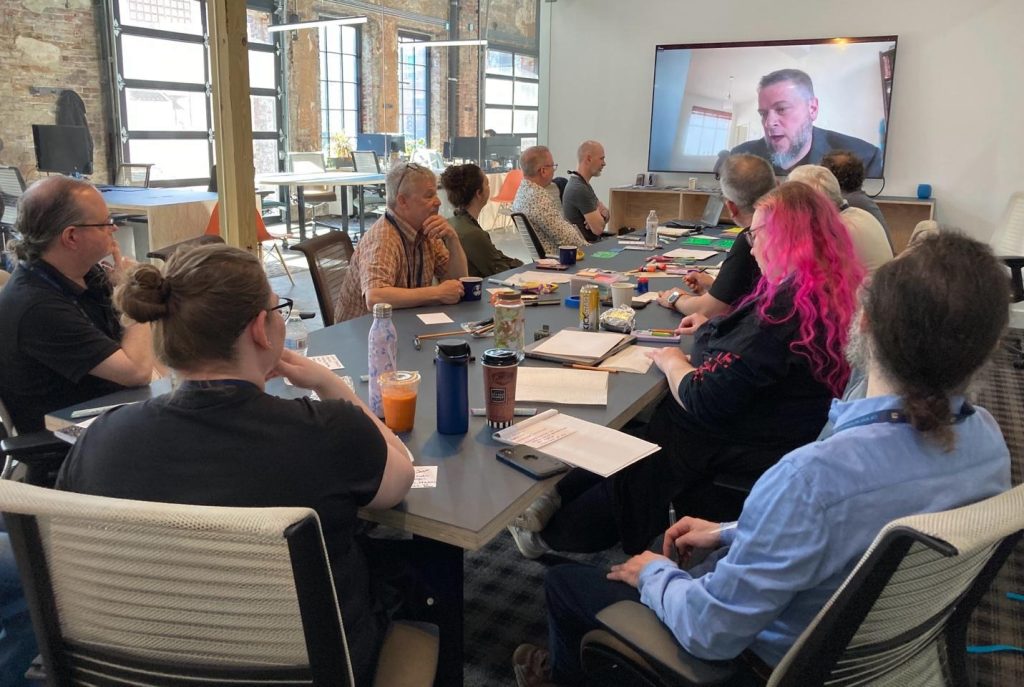 People sitting in a conference room around a table with someone on a video monitor at the front of the room.